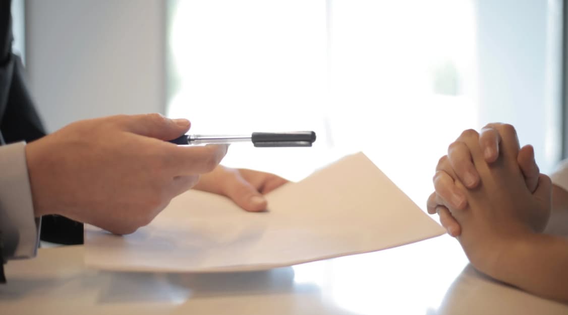 A person's hands clasped together on a table, as another person offers them a pen