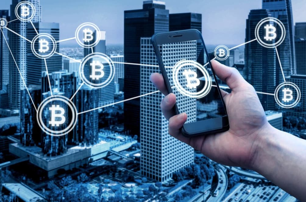 A hand holding a phone with Bitcoin symbols connected to skyscrapers
